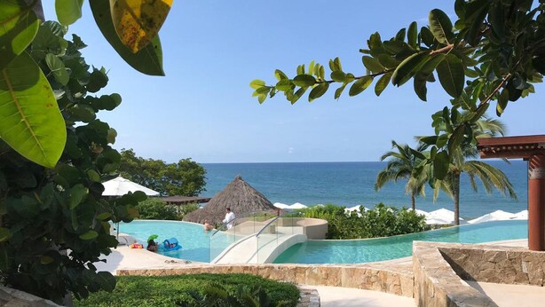 A resort pool area with a view of the ocean, surrounded by palm trees and a thatched-roof structure. - Flat 701 Luxury Condo, Premier Golf Membership, Private Pool & Golf Cart (Punta Mita)