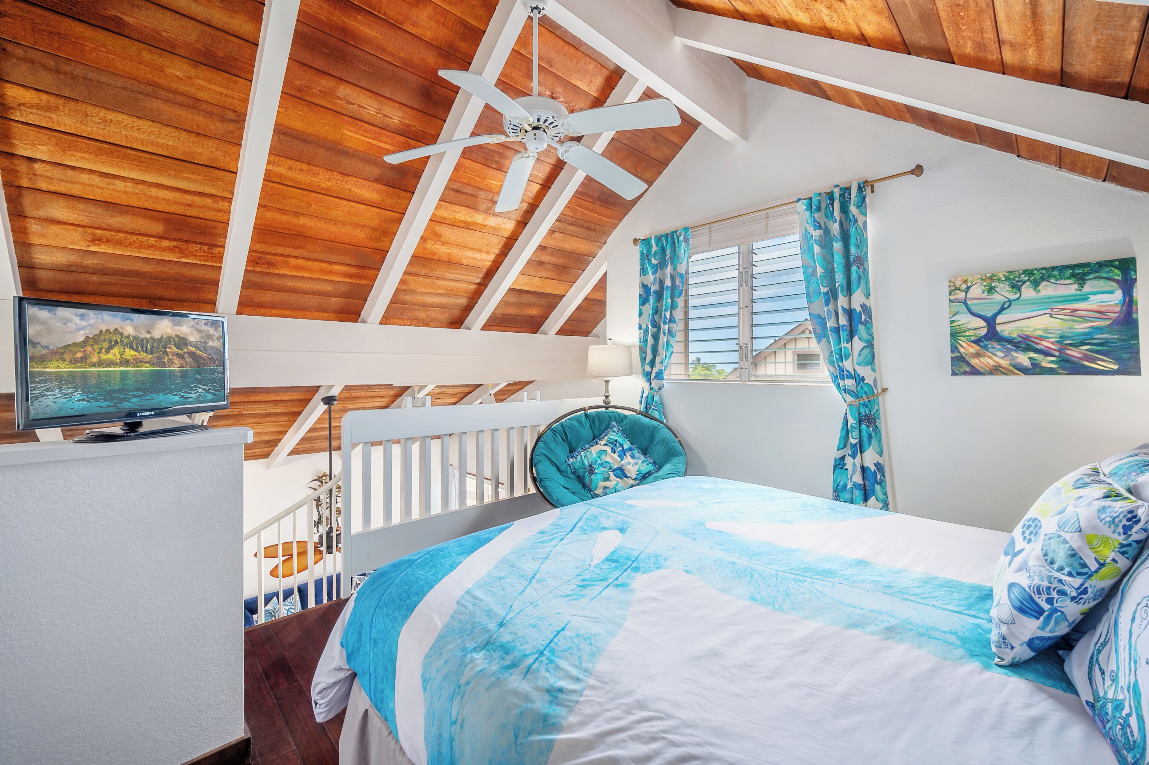 Une chambre avec un plafond en bois, un lit avec une literie bleue et blanche, un ventilateur de plafond, un téléviseur posé sur un meuble et un tableau accroché au mur.