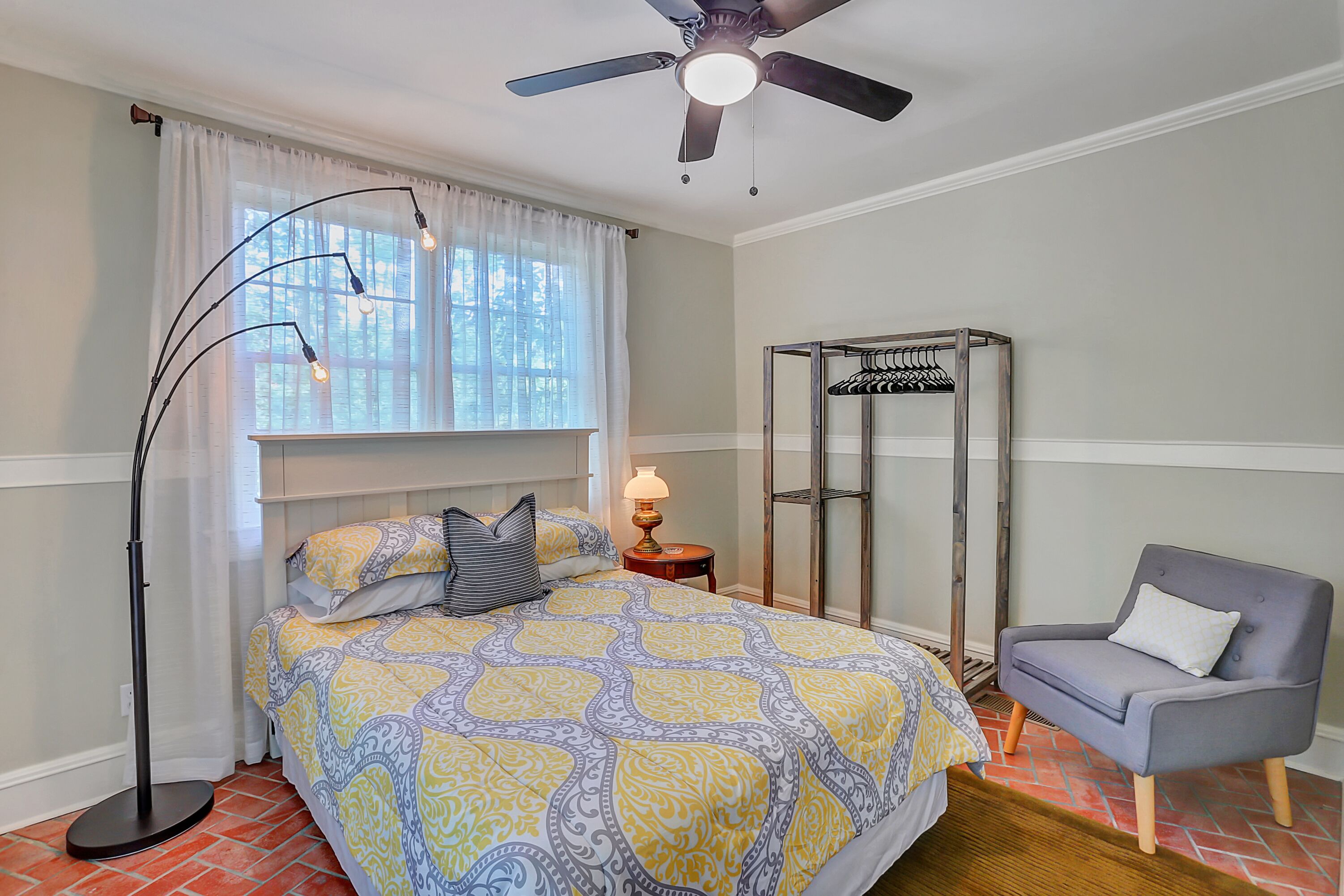 A bedroom with a bed, a chair, a floor lamp, a ceiling fan, and a window with curtains.