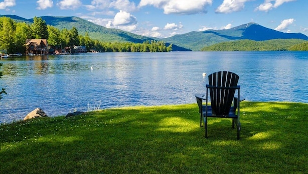 A lakeside view with a chair on the grass, a house by the water, and mountains in the background. - Family Friendly Lake Placid Condo at Prestigious Whiteface Club-Resort-Sleeps 6 (Lake Placid)