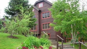 A two-story wooden house with a red trim, surrounded by lush greenery and a small garden with various plants. - Family Friendly Lake Placid Condo at Prestigious Whiteface Club-Resort-Sleeps 6 (Lake Placid)