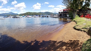 A lakeside view with boats docked, a sandy beach, and a building with outdoor seating. - Family Friendly Lake Placid Condo at Prestigious Whiteface Club-Resort-Sleeps 6 (Lake Placid)