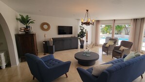 Ein geräumiges Wohnzimmer mit großem Fernseher, bequemen Sitzgelegenheiten, einem zentralen Couchtisch und Blick auf einen Poolbereich im Freien.