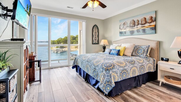 A bedroom with a large bed, a ceiling fan, a TV, and a view of the outdoors. - View for Miles! Remodeled Lands' End Luxury Condo - Wifi, Waterpark, Pool, Docks (Osage Beach)