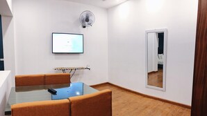 Ett rum med ett glasbord, orangefärgade stolar, en väggmonterad tv, en bordsfläkt och en spegel på väggen.