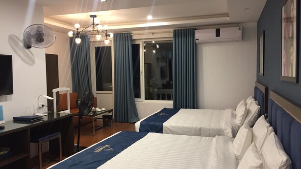 Ett hotellrum med två sängar, ett skrivbord, en stol, en skrivbordslampa, en TV som är monterad på väggen och ett fönster med gardiner.