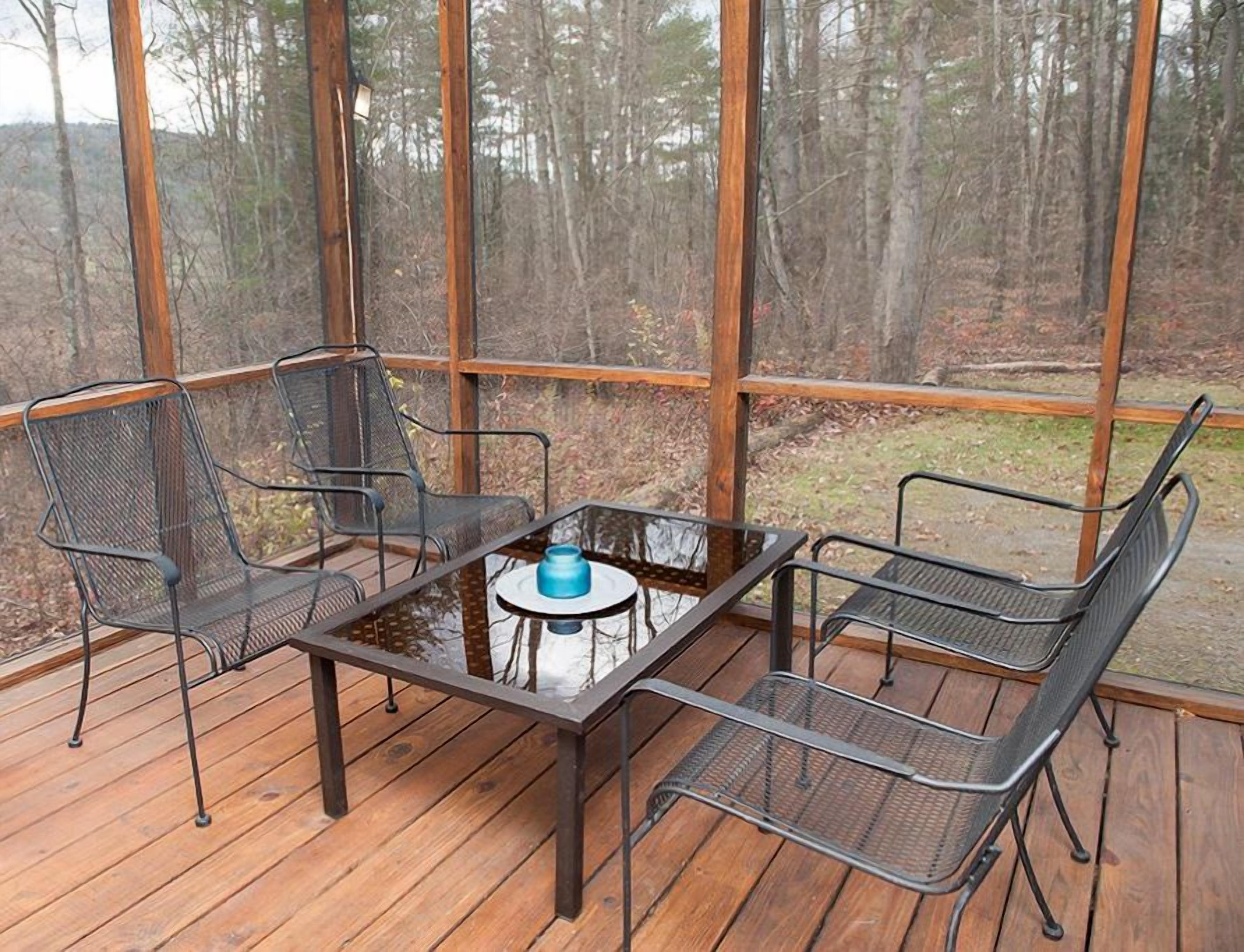 A covered outdoor seating area with a glass table and metal chairs, surrounded by a wooden deck and forest views.