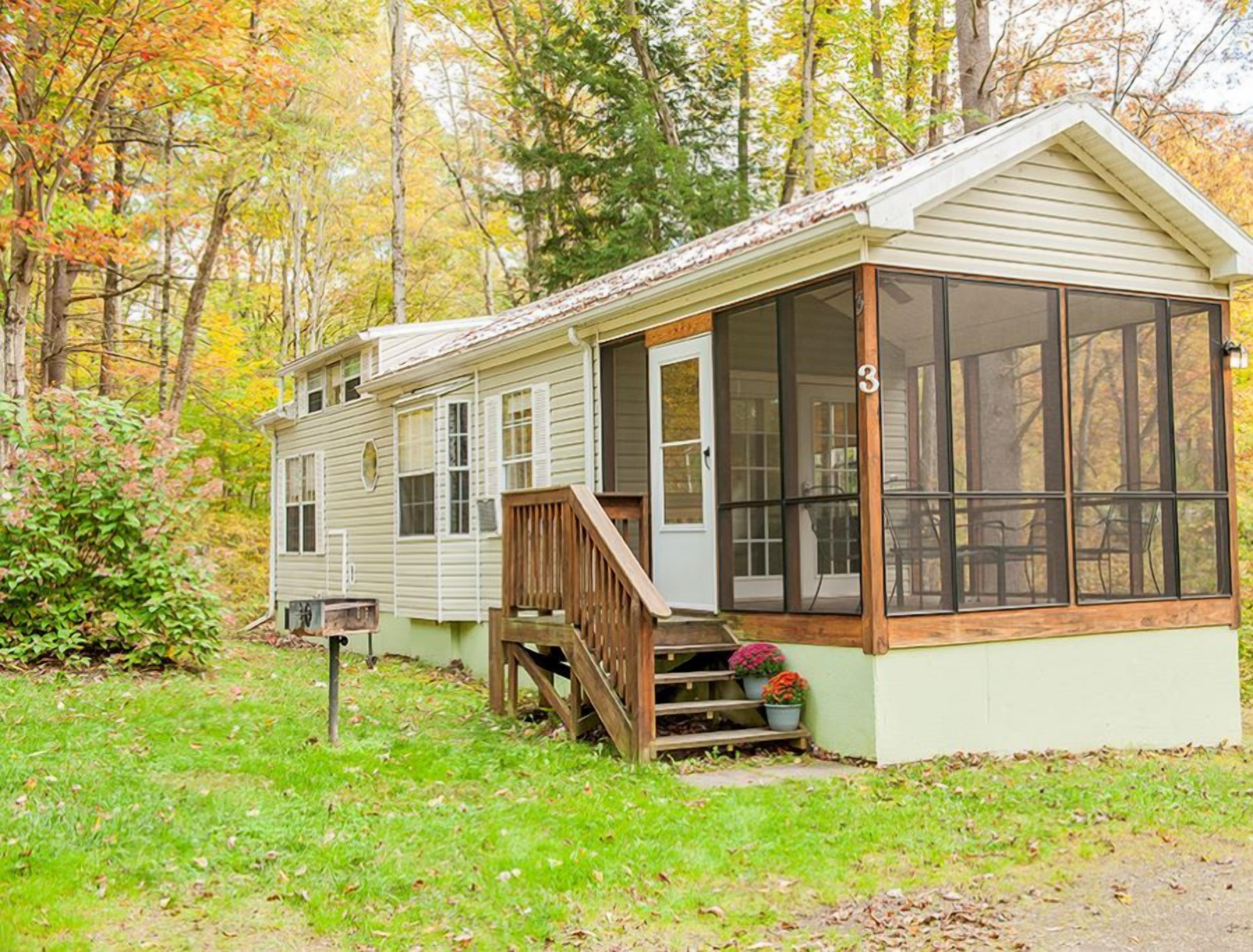 A single-story mobile home with a porch and a large screened window, surrounded by trees.