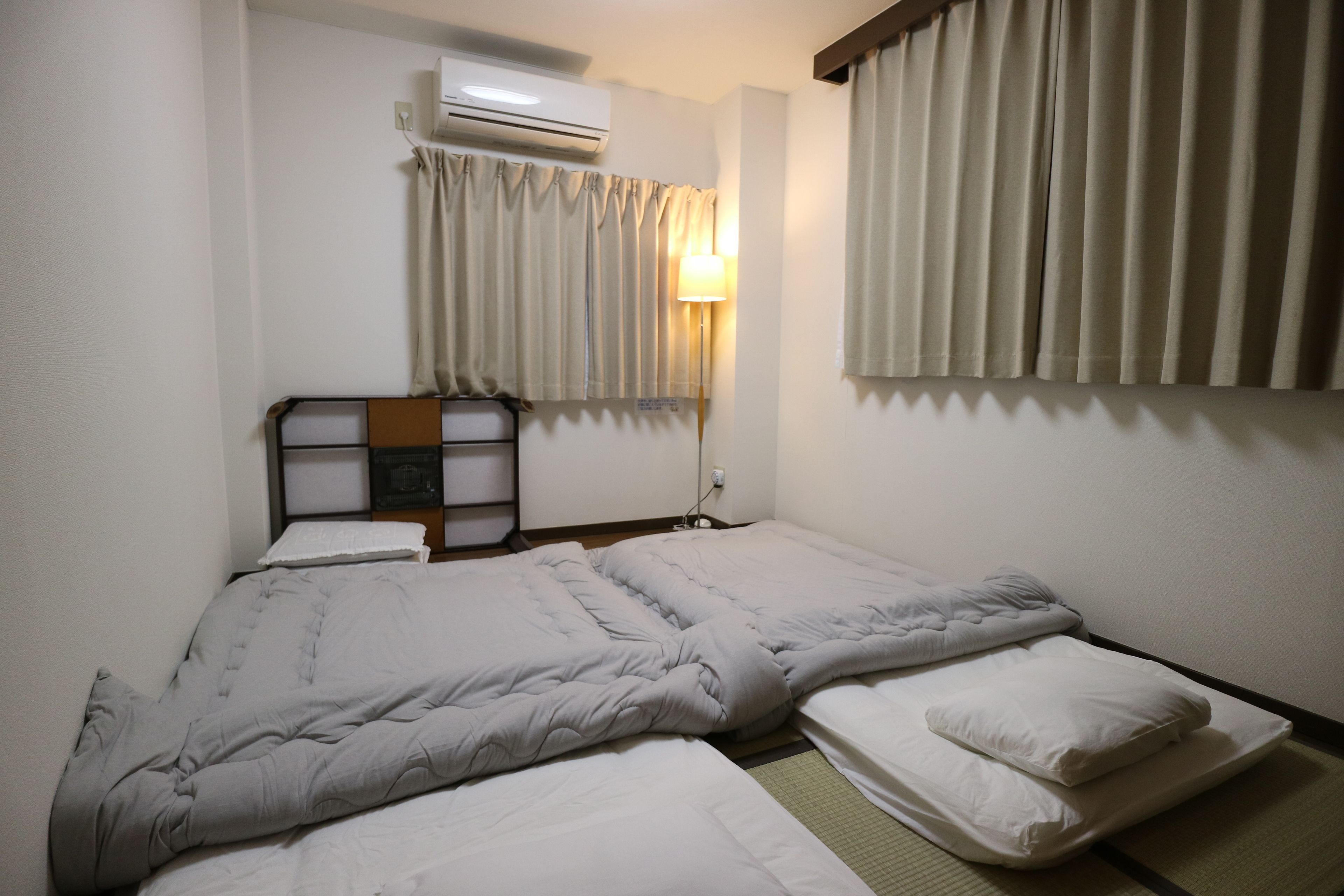 Una habitación de hotel con cama, mesita de noche, aire acondicionado y ventana con cortinas.