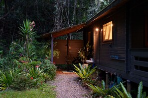 A wooden cabin with a gravel path leading to the entrance, surrounded by lush greenery and various plants.