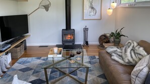 En stue med en sort brændeovn, et fjernsyn, et glas-sofabord, en brun sofa og et billede i ramme på væggen.