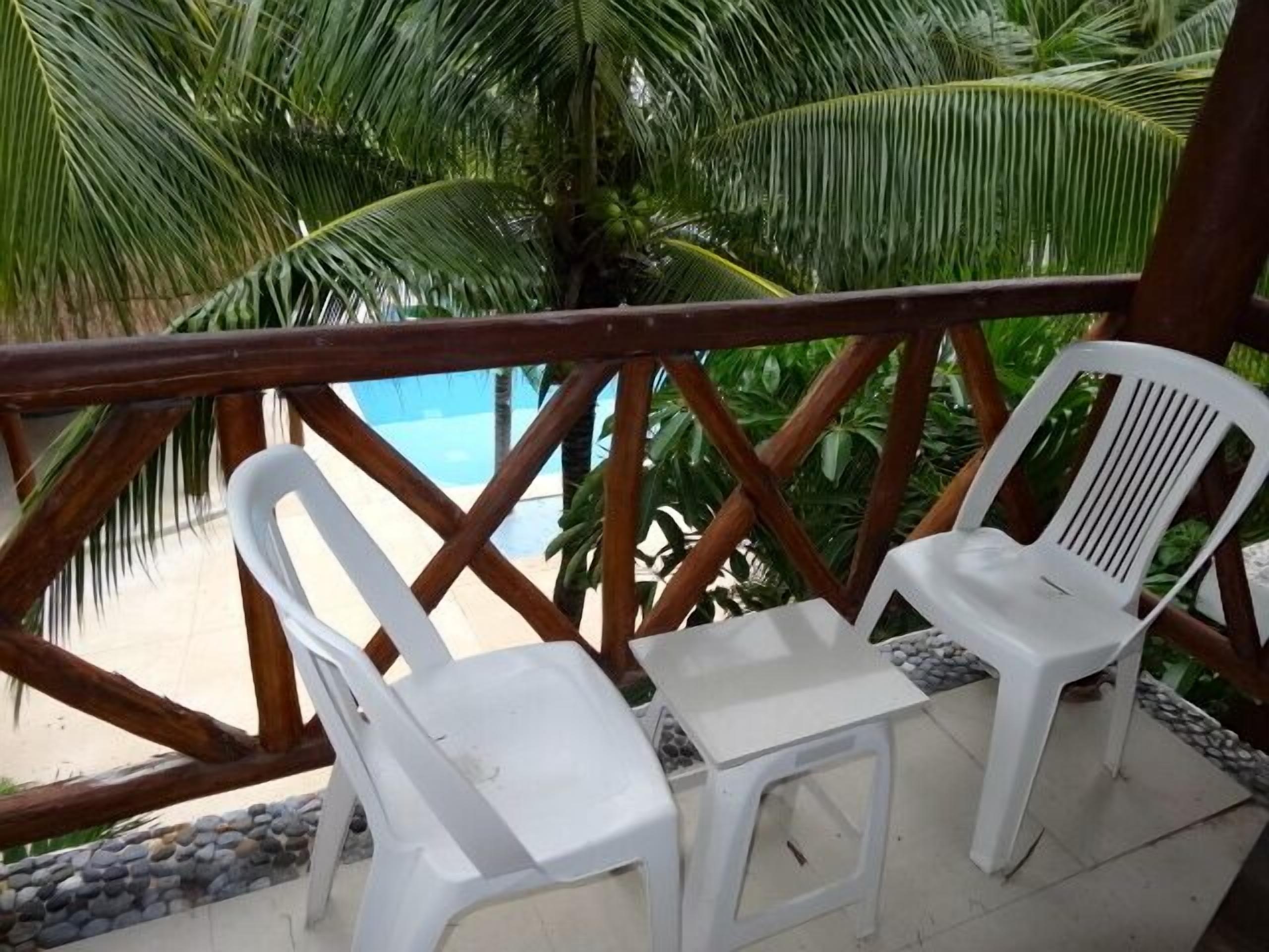 Deux chaises en plastique blanc et une petite table sont disposées sur un balcon doté d’une balustrade en bois, d’où l’on peut admirer une piscine et des palmiers.