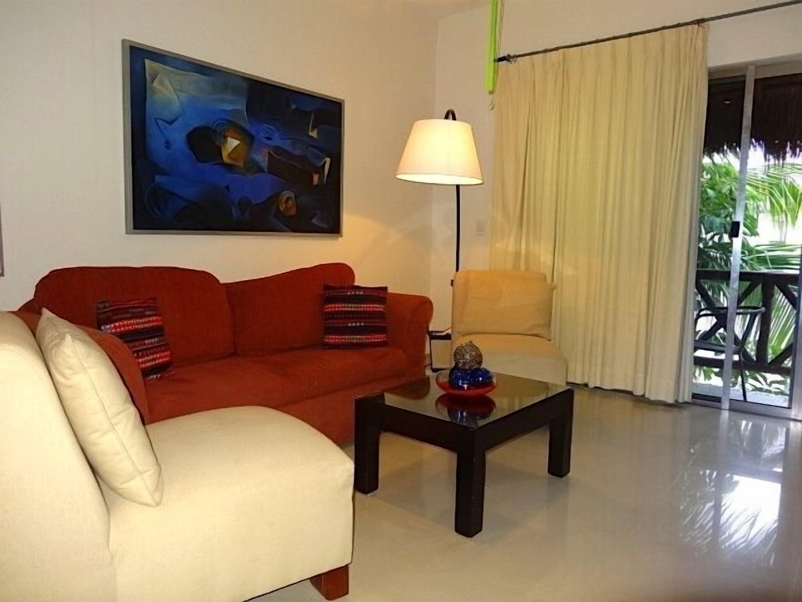 Uma sala de estar com um sofá vermelho, uma poltrona branca, uma mesa de centro de vidro e um quadro na parede.