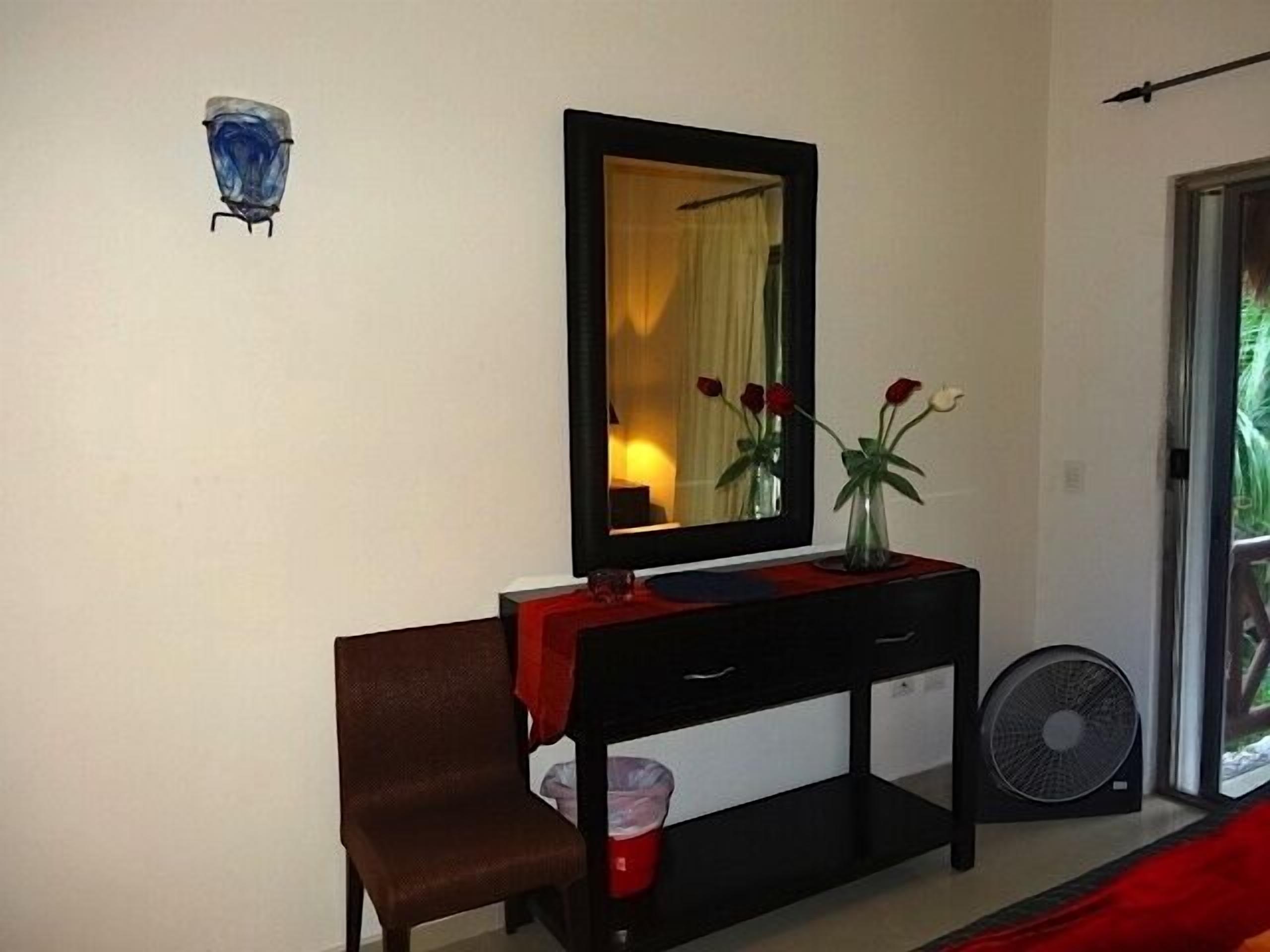 A room with a mirror, a chair, a small table with a vase of flowers, a fan, and a window with a view of greenery.