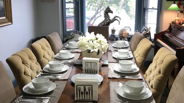 Uma sala de jantar com uma mesa comprida posta para seis pessoas, rodeada por cadeiras estofadas, e uma estátua decorativa de cavalo e um piano ao fundo.