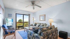 A living room with a view of the beach, a ceiling fan, a TV, and a sofa with a tropical print. - St. Augustine Oceanfront Rental – Pools, Balcony & Beach Access (St. Augustine)