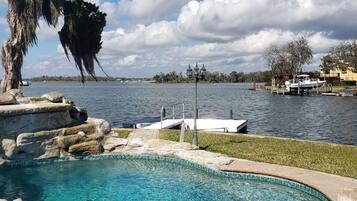 A clear swimming pool with a stone edge, a dock with a boat, and a large tree with hanging moss.