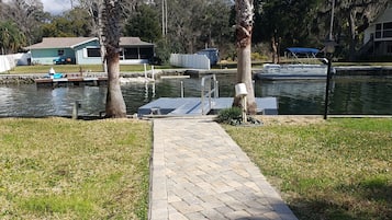 A paved walkway leading to a dock by a body of water, with palm trees and a house in the background.