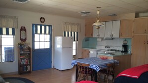 Un ambiente compatto con angolo cottura, zona pranzo con tavolo e sedie, libreria e un letto con coperte rosse e blu.