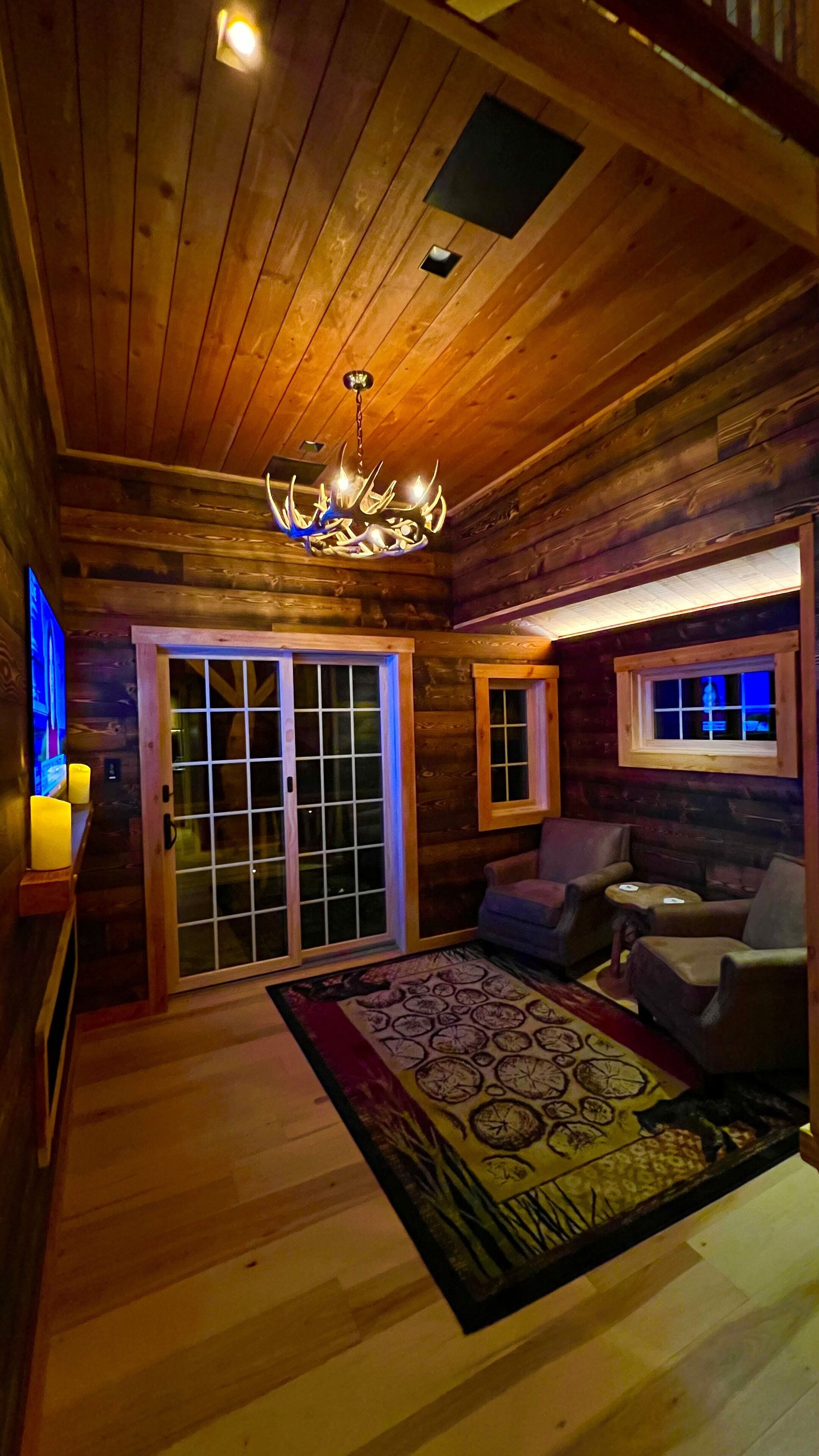 A log cabin interior with a chandelier, wooden walls, a sliding glass door, and a patterned rug.