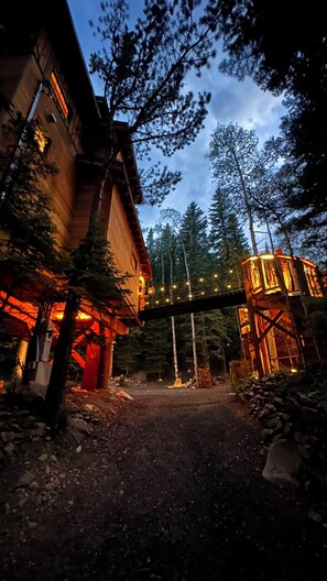 A rustic wooden building with a covered outdoor area, surrounded by tall trees and a wooden bridge overhead. - Ultimate Cabin and Treehouse experience - just 1 hour from Denver. (Black Hawk)