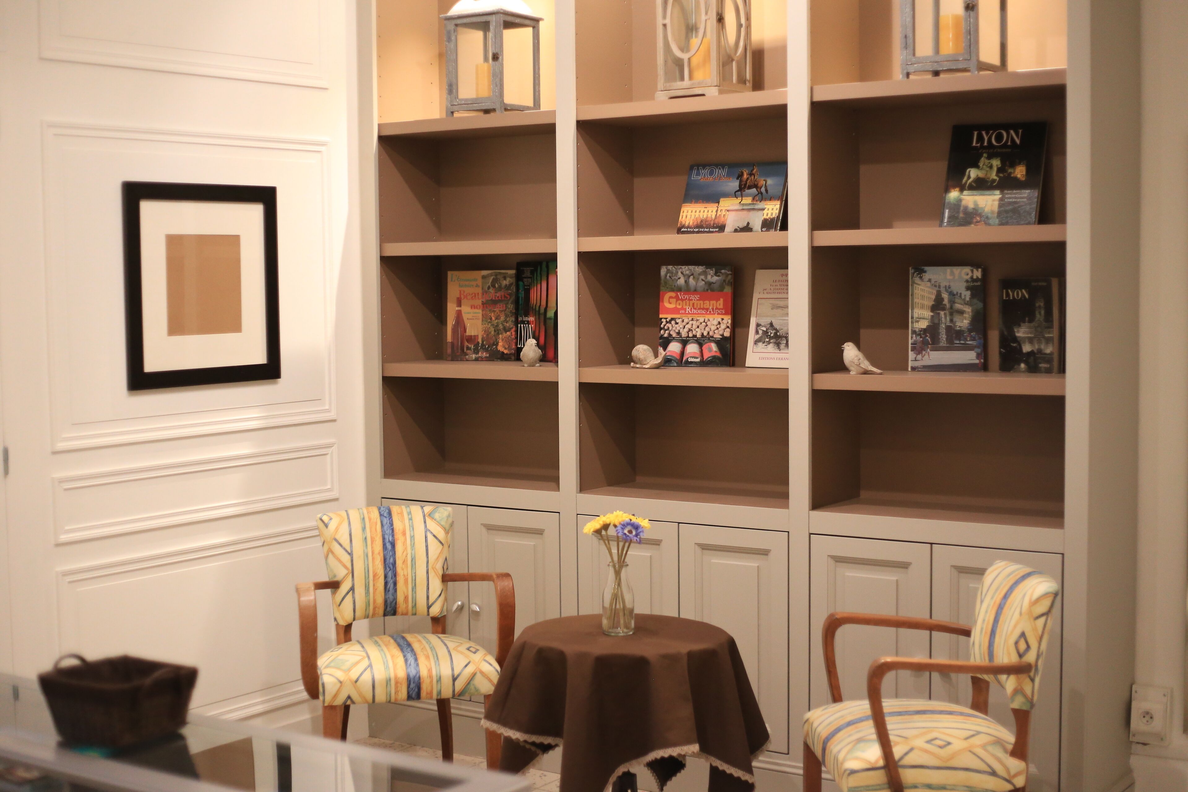 A room with a bookshelf, two patterned chairs, a small table with a vase, and a framed picture on the wall.
