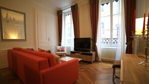 A living room with a red sofa, a TV on a wooden stand, and a window with curtains. - 3 bedrooms Premium Apartment - La Suite Lanterne (Lyon)