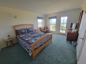 Un dormitorio con cama de madera, una mesita de noche pequeña, una silla, un escritorio con televisor y ventanas amplias con vistas.