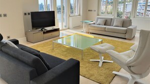 Una sala de estar moderna con un televisor de pantalla plana, una mesa de centro de vidrio, un sofá, un sillón blanco y ventanas amplias que ofrecen vistas a la ciudad.