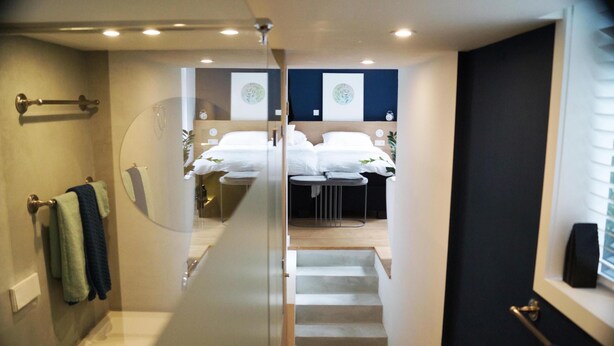 Une chambre d’hôtel moderne avec une cloison en verre, un escalier menant à une chambre à coucher équipée d’un lit et d’une petite table, ainsi qu’une salle de bain avec des toilettes et un porte-serviettes.
