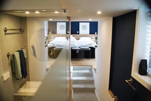 A modern hotel room with a glass partition, a staircase leading to a bedroom with a bed and a small table, and a bathroom with a toilet and towel rack.