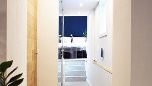Un couloir étroit, avec une porte en bois sur la gauche, un tapis à motifs sur le sol et un escalier menant à une pièce où se trouvent un lit et une plante.