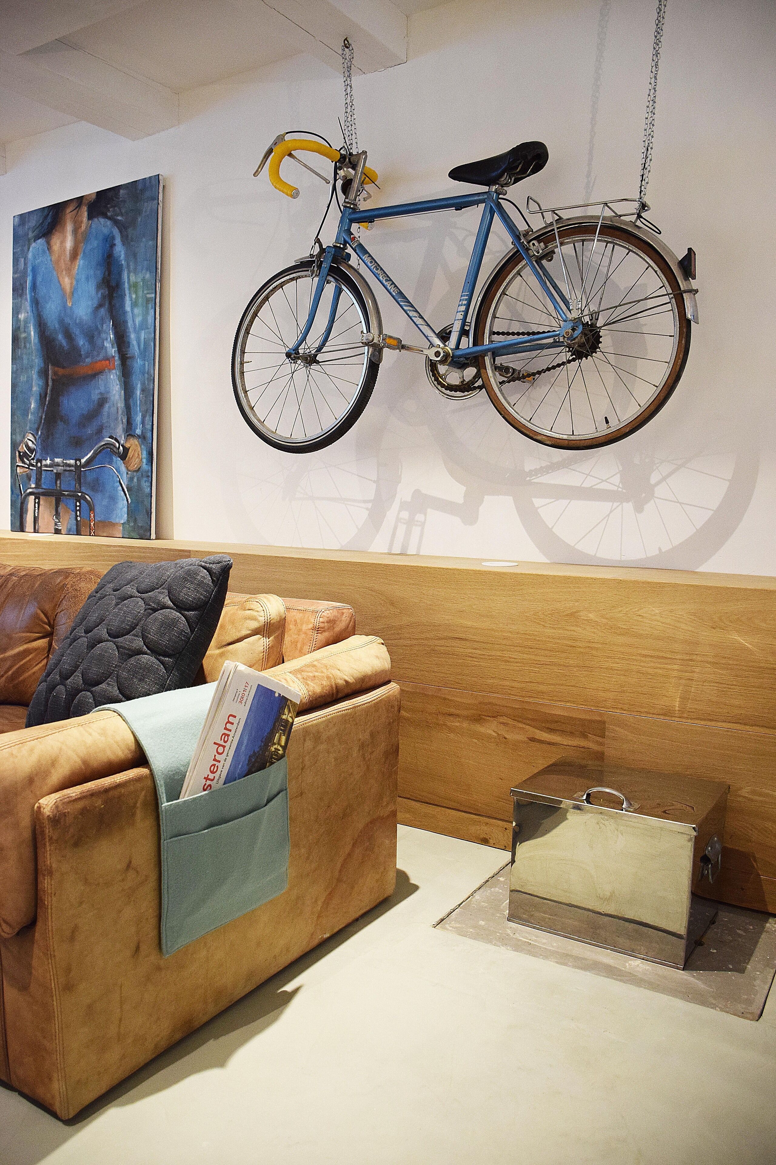 A room with a brown leather sofa, a blue bicycle hanging from the ceiling, and a painting of a bicycle on the wall.