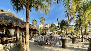 Un restaurant situé en bord de mer, doté d’une terrasse, de palmiers et d’une structure surmontée d’un toit de chaume.