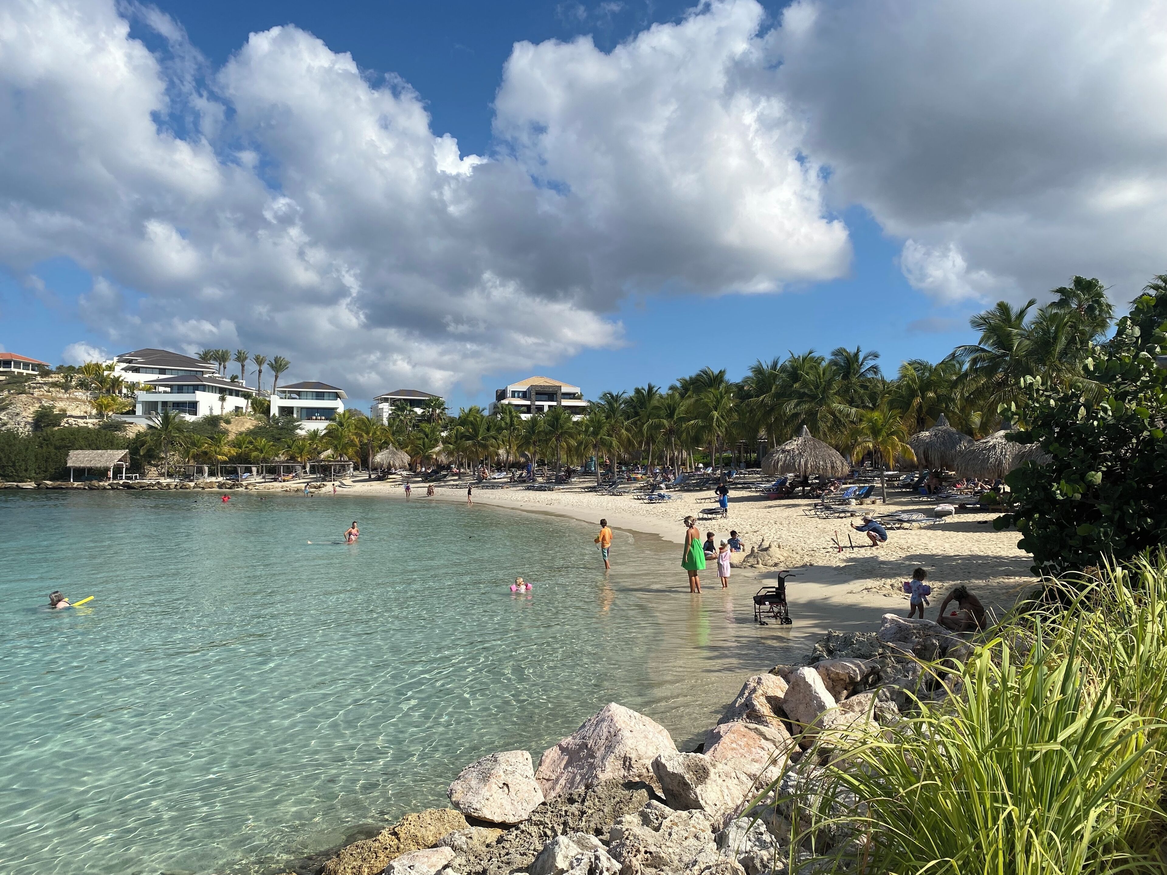 Une plage à l’eau cristalline, bordée de palmiers, avec des bâtiments modernes en arrière-plan.