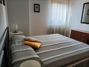 Una cama bien hecha con ropa de cama a rayas, una mesita de noche con una lámpara, un aparador de madera y una ventana con cortinas traslúcidas.