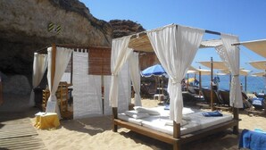Una cabaña en la playa con una cama de madera y dosel blanco, rodeada de sillas y sombrillas.
