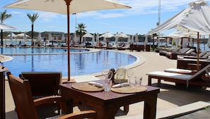 Zona junto a la piscina con sillas de descanso, sombrillas y una mesa con vasos y una botella.