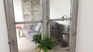 Ein rustikaler Spiegel mit verwitterter Rahmen, der einen Raum mit einem grauen Sofa, einem kleinen Tisch und einer hängenden Pflanze zeigt.