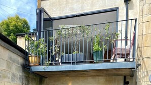 Un edificio de paredes de piedra con una puerta oscura, una ventana pequeña y un balcón con macetas.