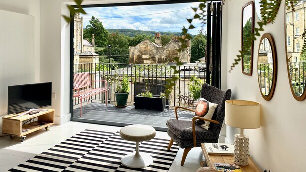 Una sala de estar moderna con un televisor grande, un sillón cómodo, una mesita y vistas a un paisaje urbano a través de una puerta de vidrio.