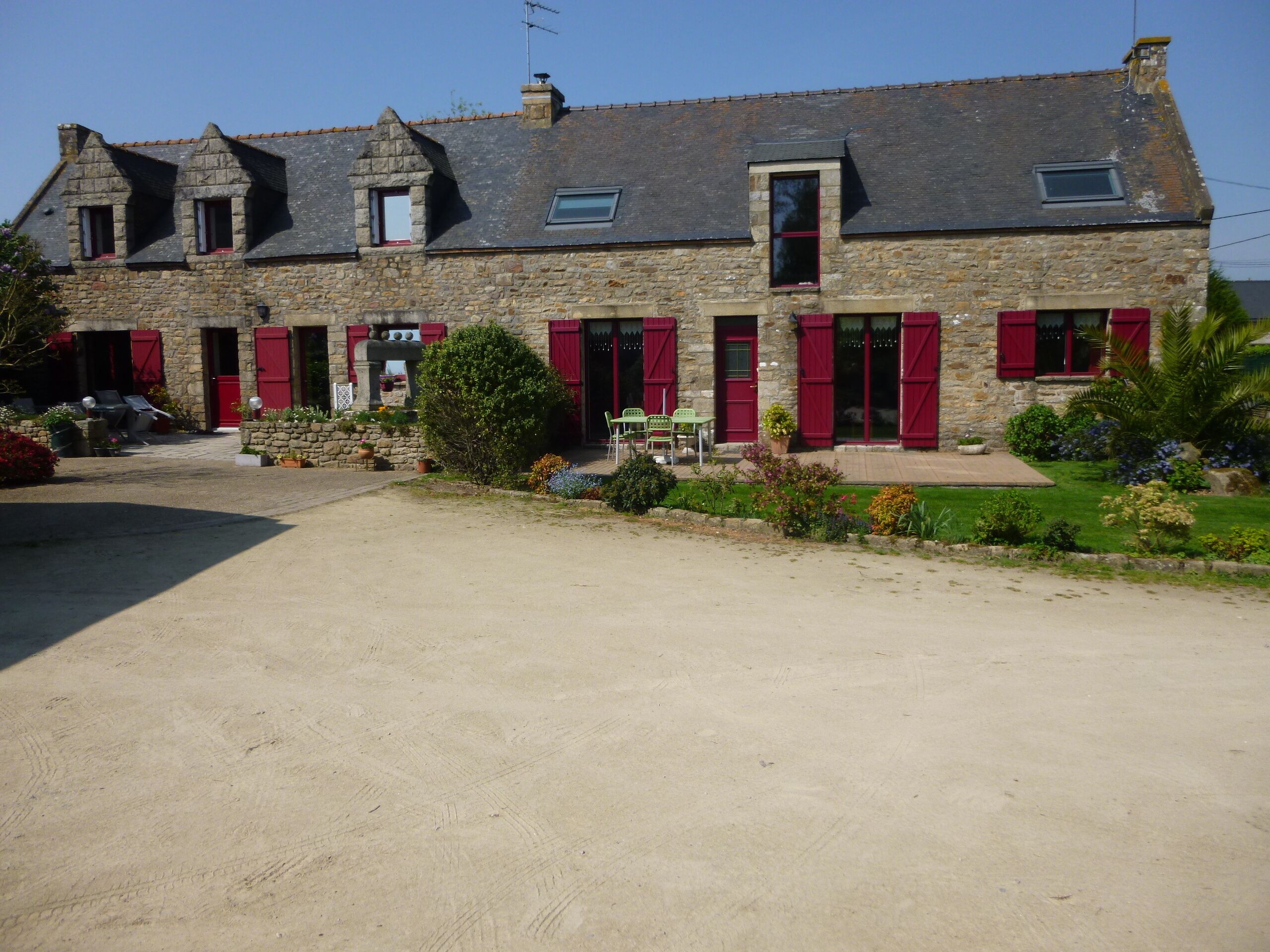 A stone building with red shutters, a paved driveway, and a small garden with chairs and plants.