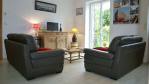 Ein Wohnzimmer mit zwei Ledersofas, einem Holztisch, einem Fernseher auf einem Schrank und einem großen Fenster mit Blick auf Grünflächen.