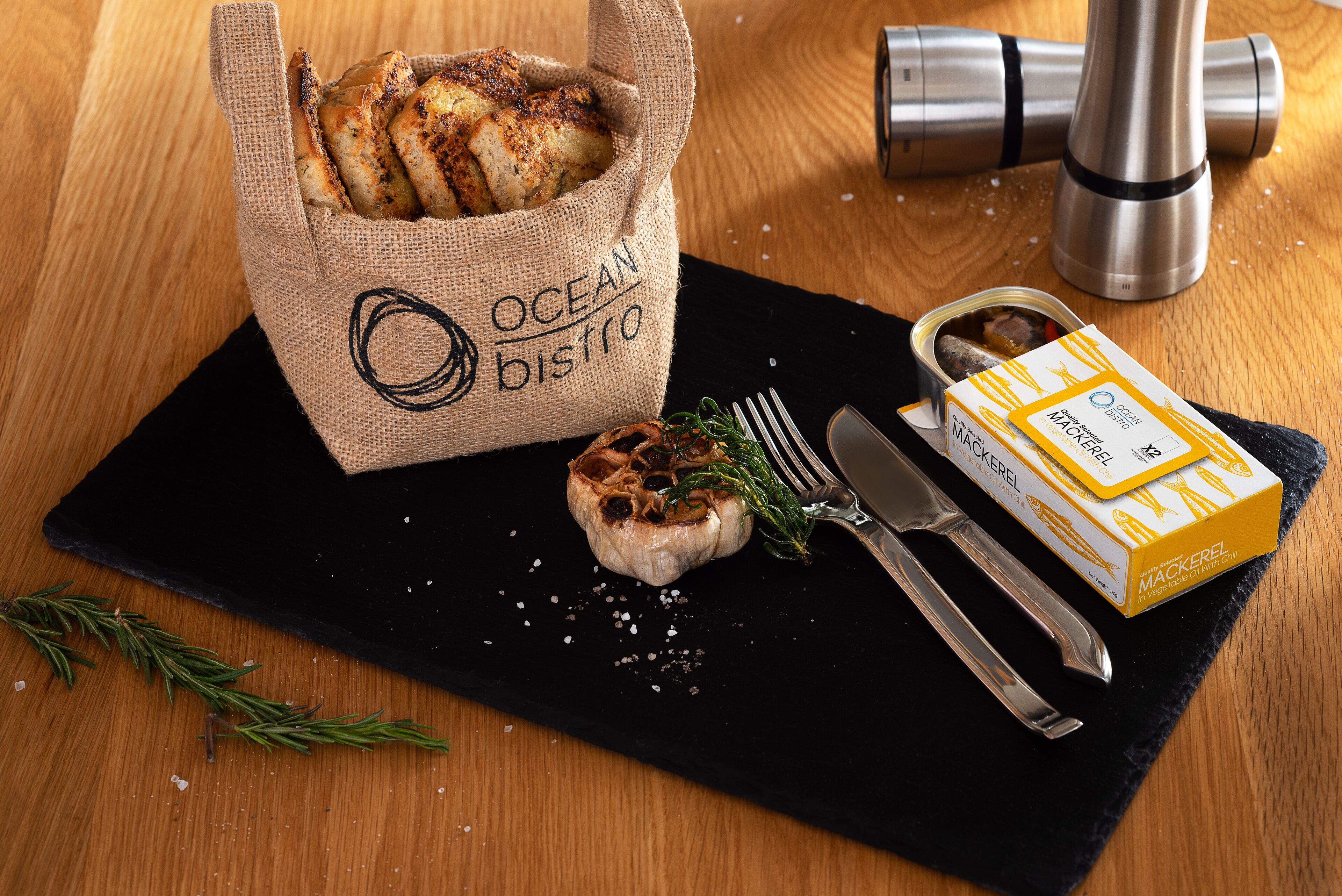 Una bolsa de arpillera con la etiqueta "OCEAN DISTRO" que contiene pan, un diente de ajo asado con hierbas, una caja de boquerones y un molinillo de pimienta sobre una mesa de madera.