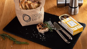 Una bolsa de arpillera con la etiqueta "OCEAN DISTRO" que contiene pan, un diente de ajo asado con hierbas, una caja de boquerones y un molinillo de pimienta sobre una mesa de madera.