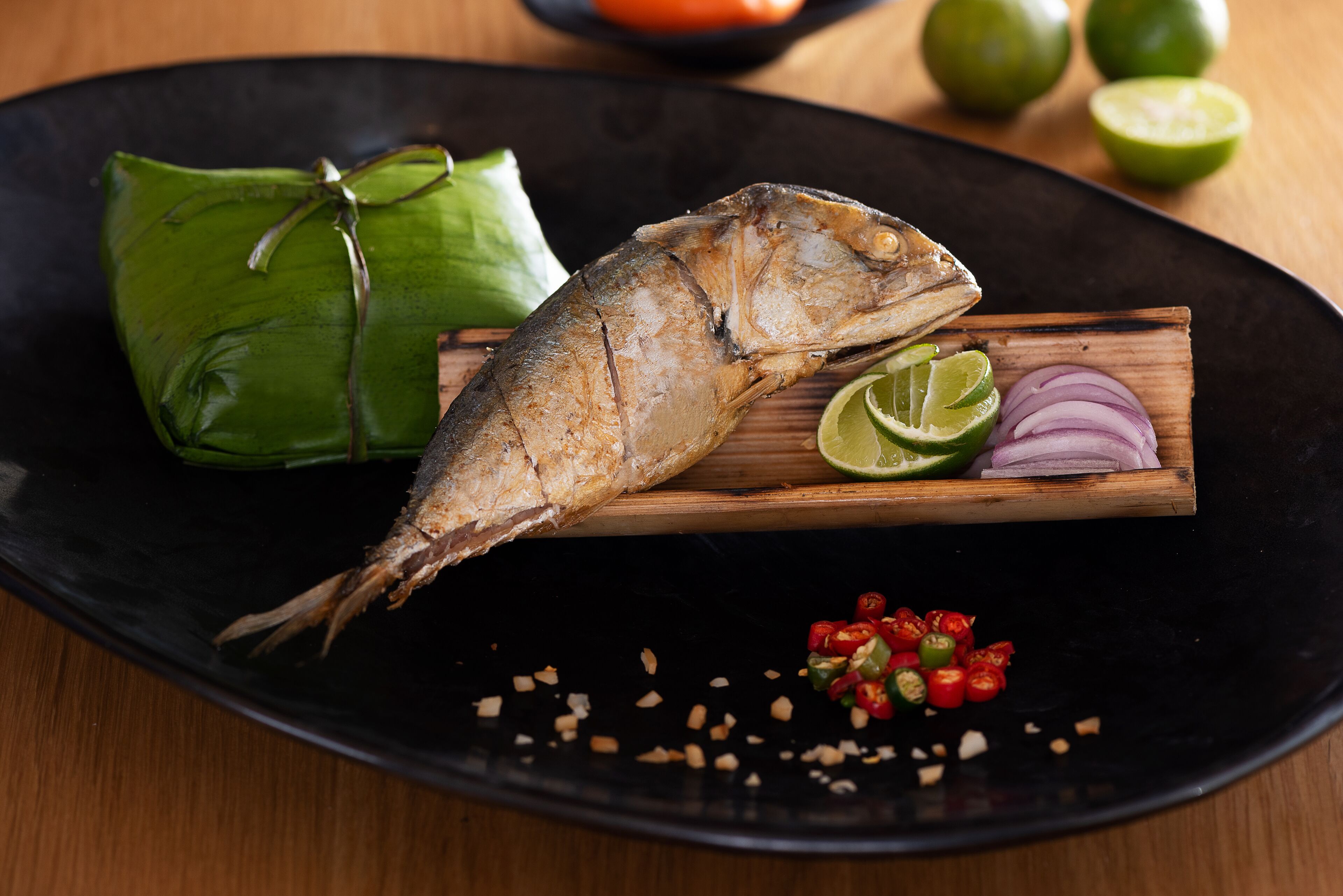 Ein Fisch auf einem Holzboden, garniert mit Limetten- und roten Zwiebelscheiben, begleitet von einem in grüne Blätter gewickelten Fisch.