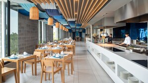 A modern dining area with wooden tables and chairs, a long counter with a chef preparing food, and large windows providing views of the outdoors. - Oceanphere Pattaya Villa (Sattahip)