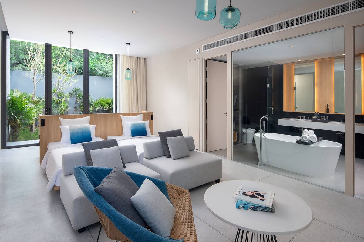 A modern bedroom with a large bed, a sofa, a wicker chair, and a round coffee table. There is a bathroom with a bathtub and a large mirror.