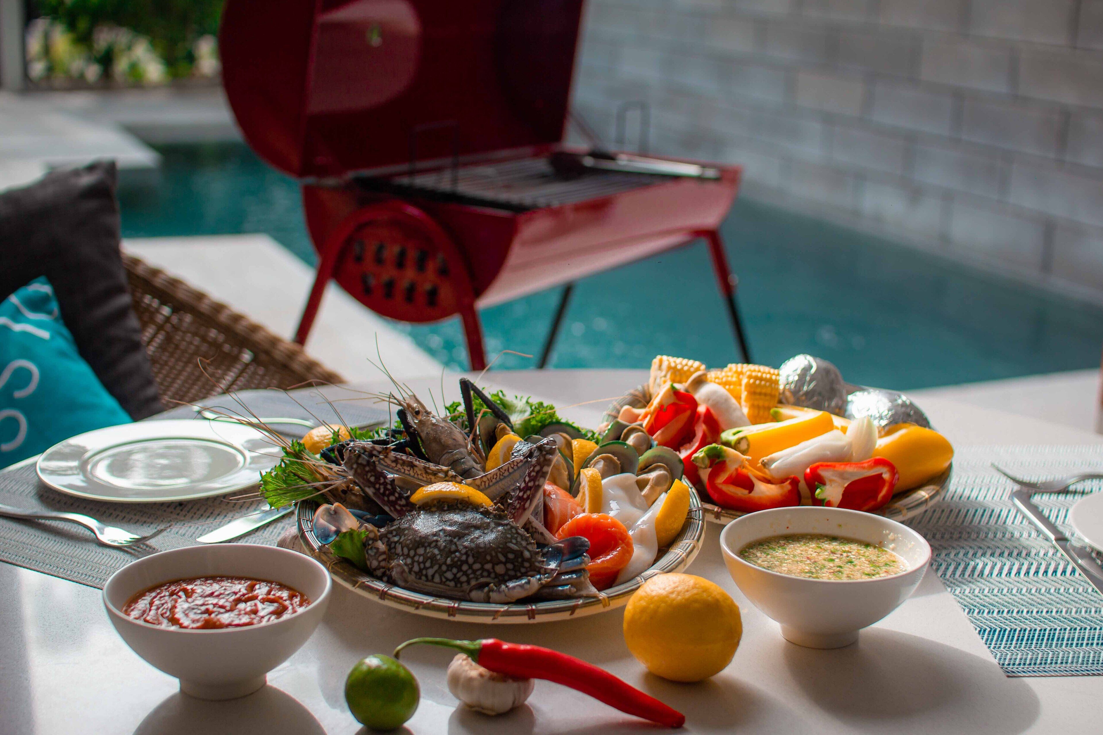 A dining table with seafood, vegetables, and condiments, near a pool and a red chair.