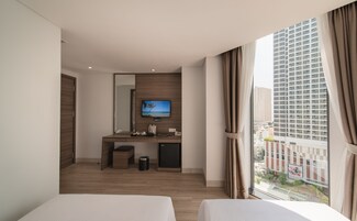 Habitación de hotel con dos camas, un escritorio con televisor, un cubo de basura y una ventana con vistas a la ciudad.